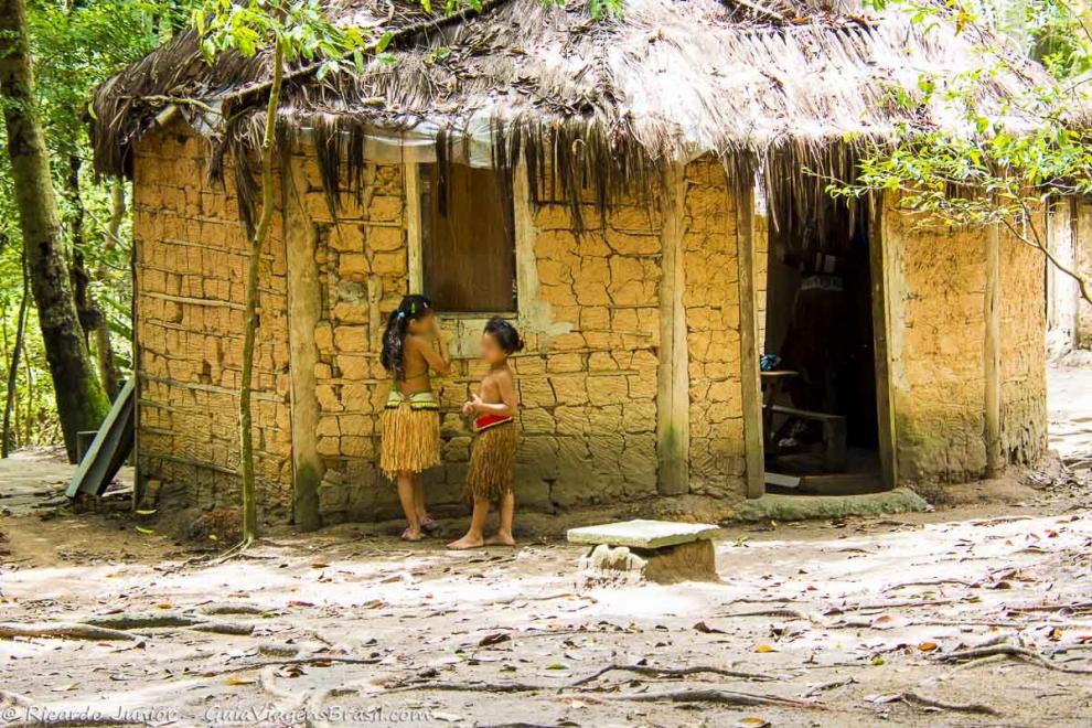 Imagem de duas meninas indígenas em frente a uma casa de barro.